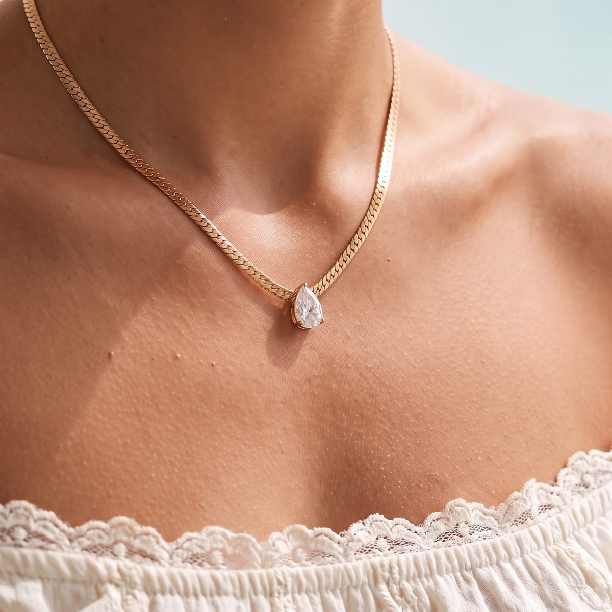 Close-up of the Pear Necklace in rose gold, a pear crystal on a flat herringbone chain above a lace top