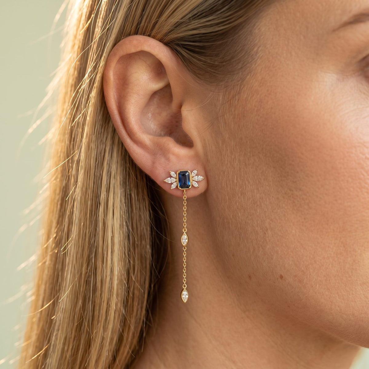 Woman wearing the Isabella Noir Drop Earrings on the lobe with a blue stone and hanging gold chain