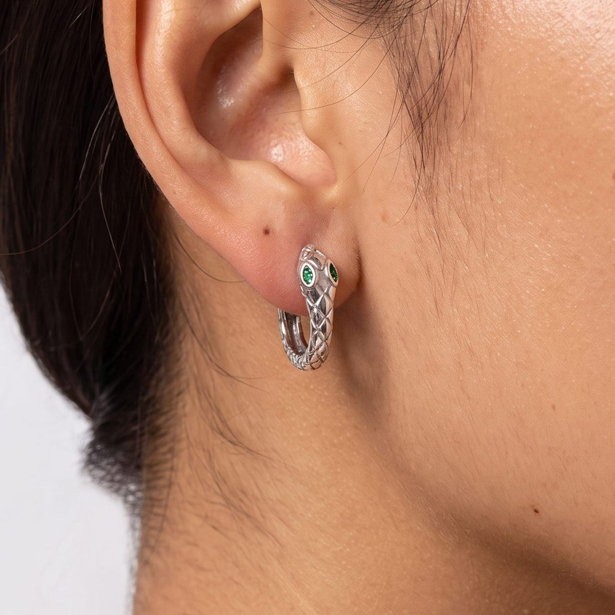 Close view of a woman wearing the Lilou Silver Snake Hoops through the lobe, featuring green stone eyes