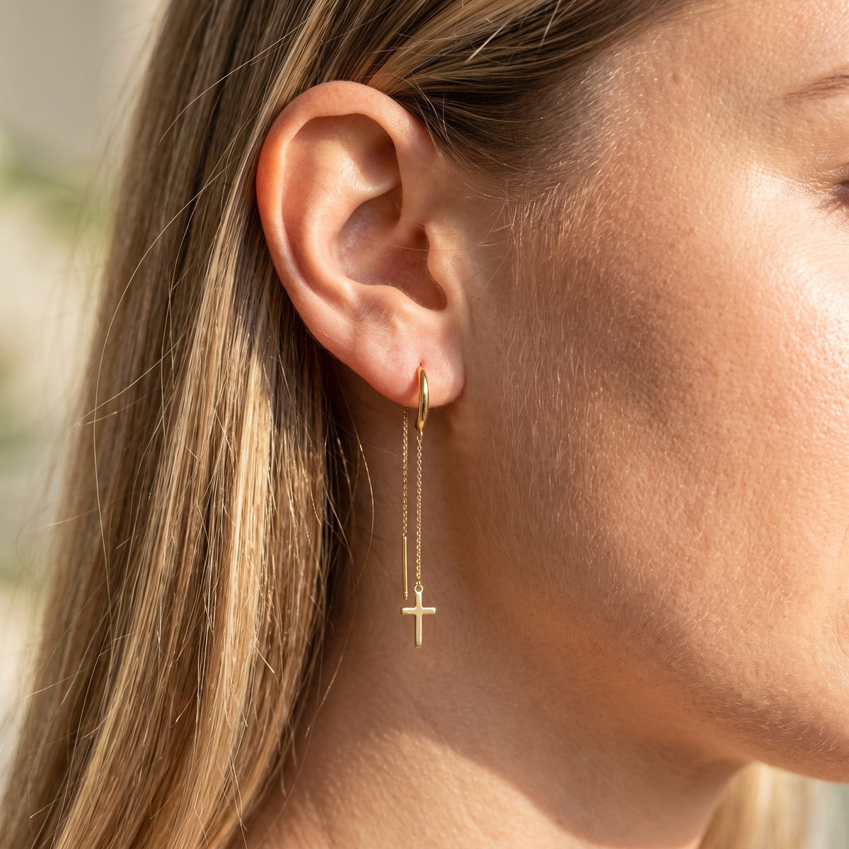 Close view of a woman wearing the Camille Cross Chain Earrings on the lobe with a dangling gold cross
