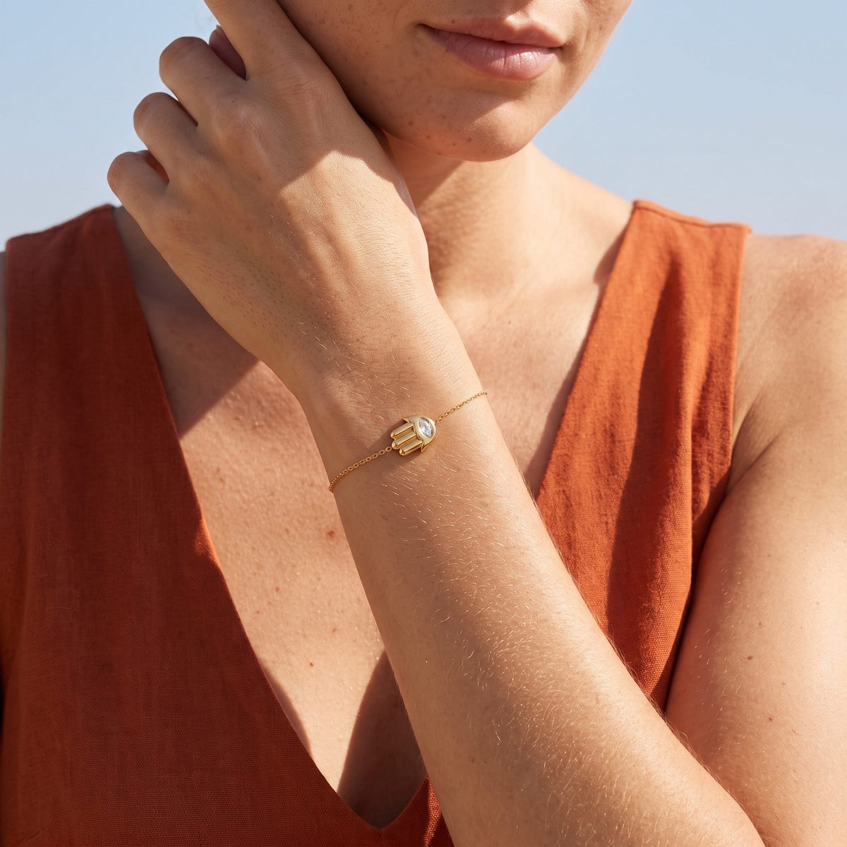 Woman wearing the Hamsa Charm Bracelet on the wrist, touching her jawline in an orange top
