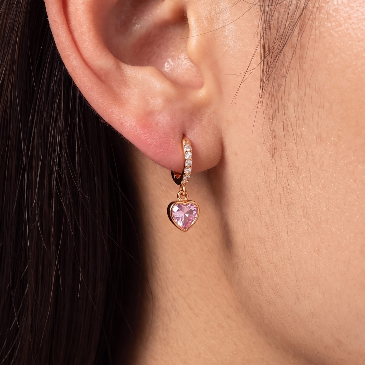 Close-up of the Pink Heart Huggies on an ear, featuring a dangling pink heart crystal charm, in rose gold