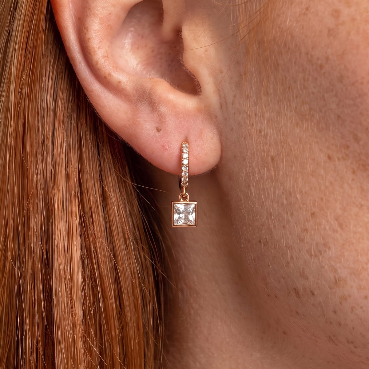 Close-up of the Square Crystal Huggies on the ear, featuring a dangling square crystal charm, in rose gold